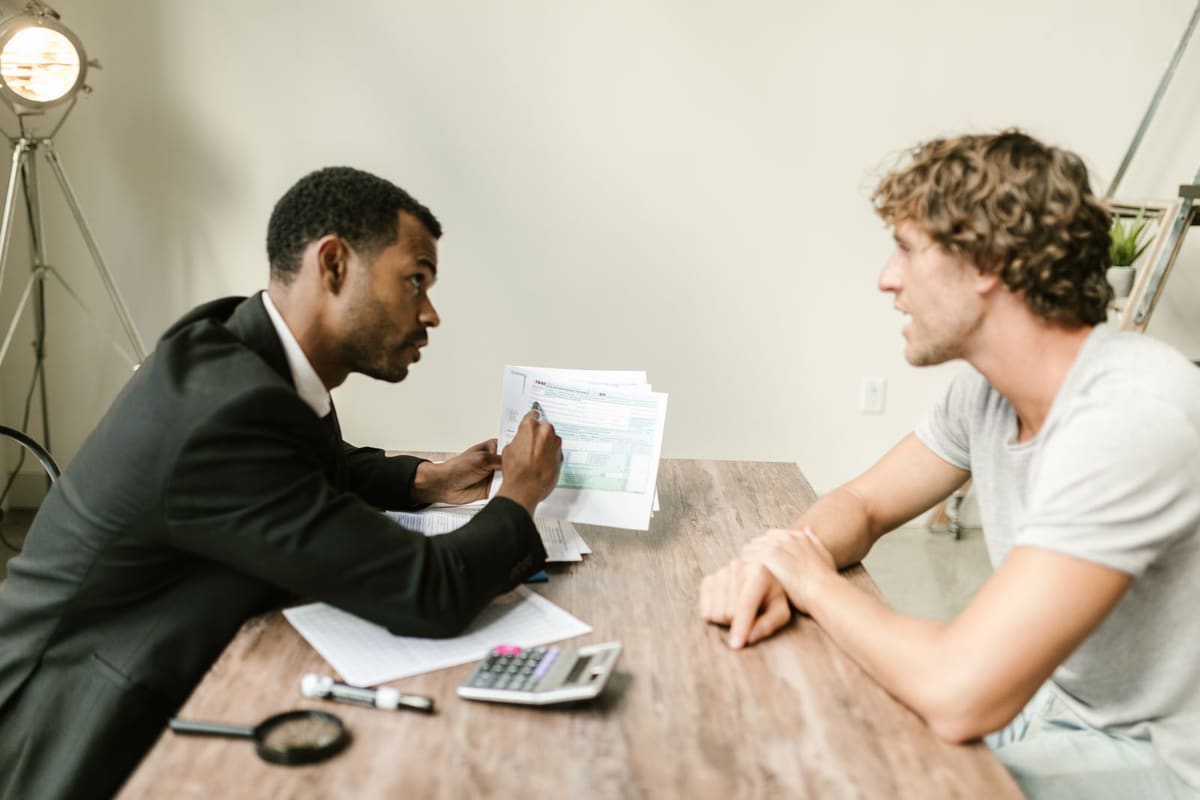 An adviser in a suit holds up a document across a table from a client, with a calculator and magnifier on the table