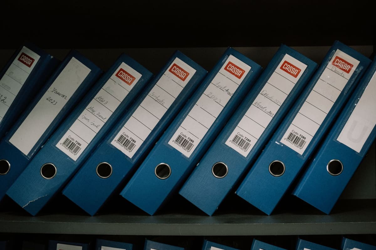 A row of dark blue lever-arch binders on a shelf, each with a white spine label showing handwritten dates