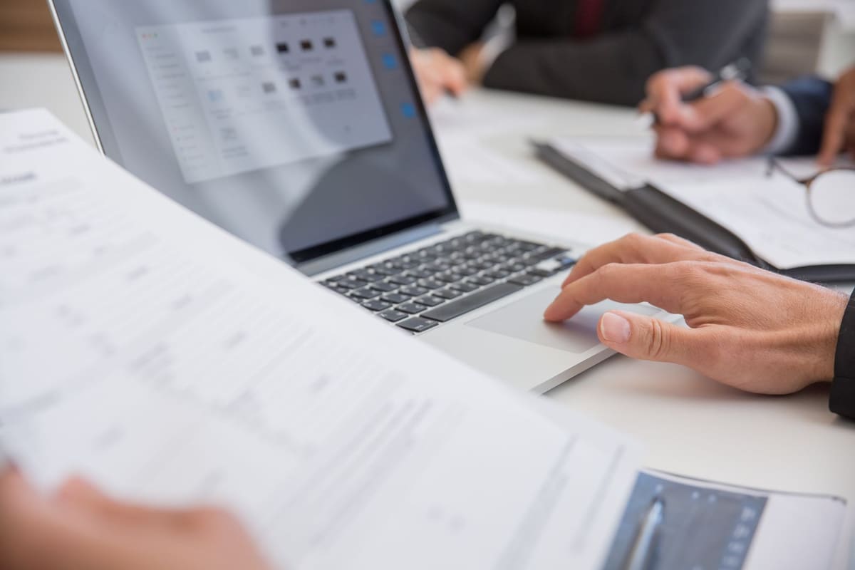 Hands typing on a laptop on a table covered with printed spreadsheets, with another person writing in the background