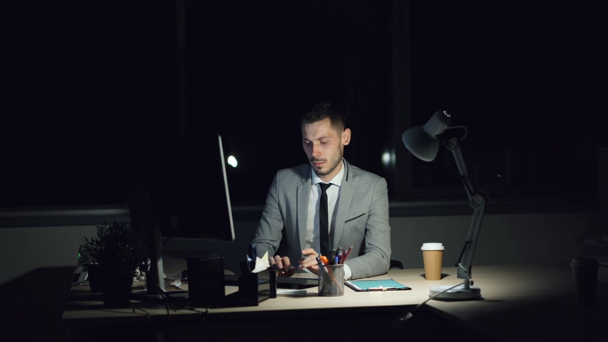 A man in a suit sits alone at a desk in a darkened office, lit only by a desk lamp and a monitor