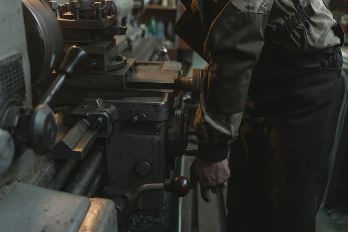 Close-up of a machinist in a worn work jacket, hand on the feed lever of a metal lathe in a workshop