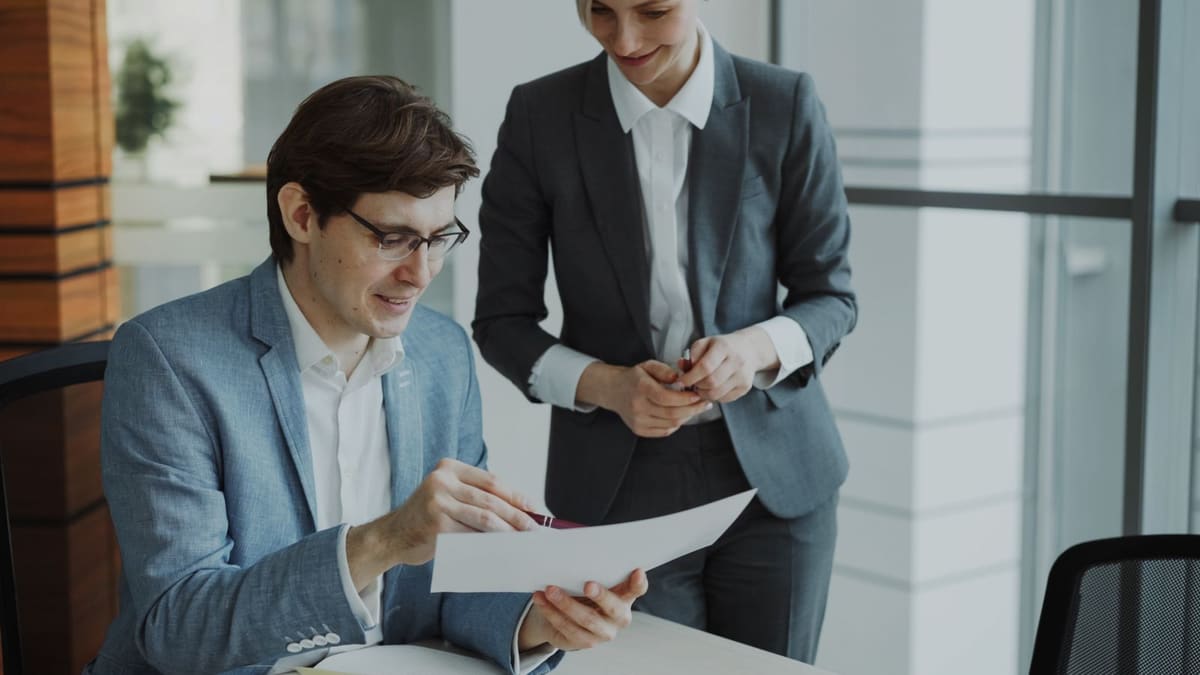 A seated man in a jacket reads a printed document while a woman in a suit stands beside him looking at the same page