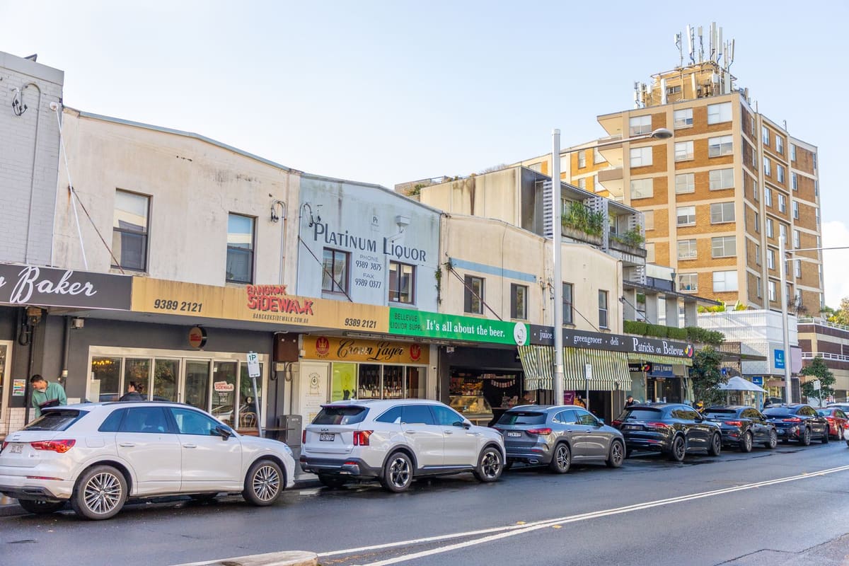 A row of suburban Sydney shopfronts in sunshine — a baker, a Thai restaurant and a liquor store under awnings, with cars parked along the kerb