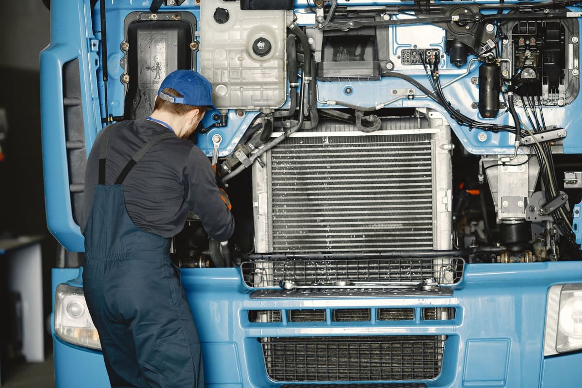 A mechanic in overalls working on the exposed radiator and engine bay of a blue truck with its front panel tilted open